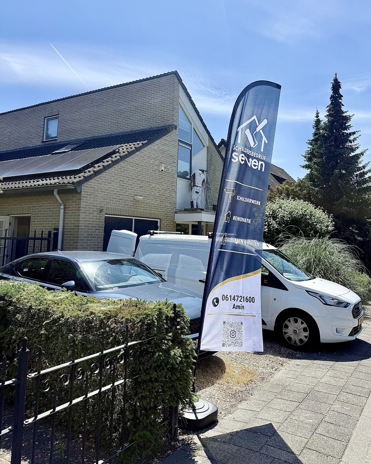 Witte bedrijfsbus met de navy beachflag van Schildersbedrijf Seven voor een woning, een schilder op de ladder op de achtergrond