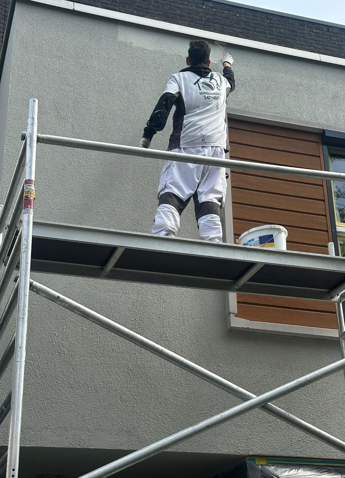 Schilder in witte overall op een steiger, bezig met het schilderen van een buitenmuur